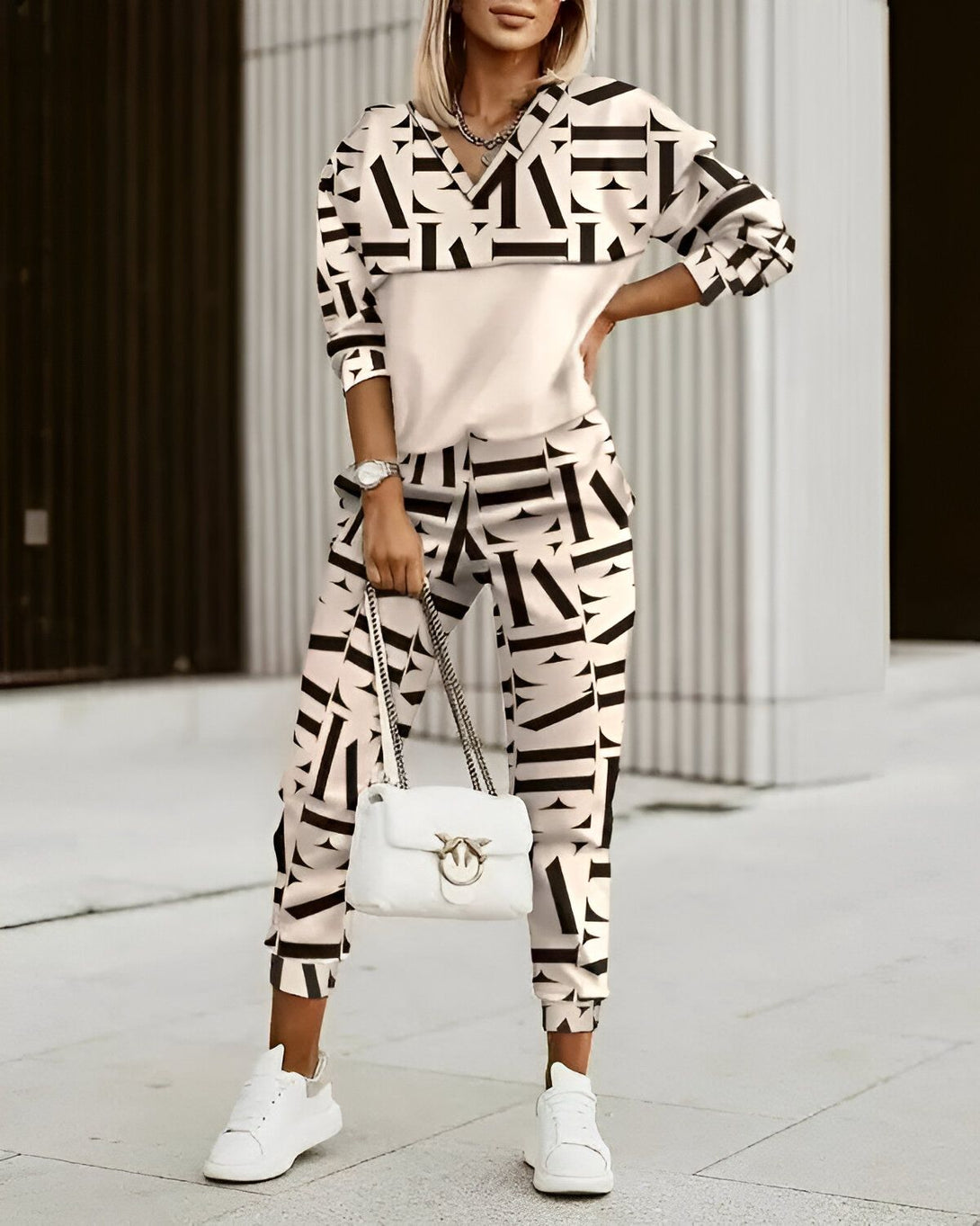 Une femme porte un survêtement beige et noir. Le survêtement est composé d'un haut à manches longues et d'un pantalon. Le haut est court et possède une encolure en V. Le pantalon est long et possède une taille élastique. La femme porte également des baskets blanches et un sac à main blanc.
