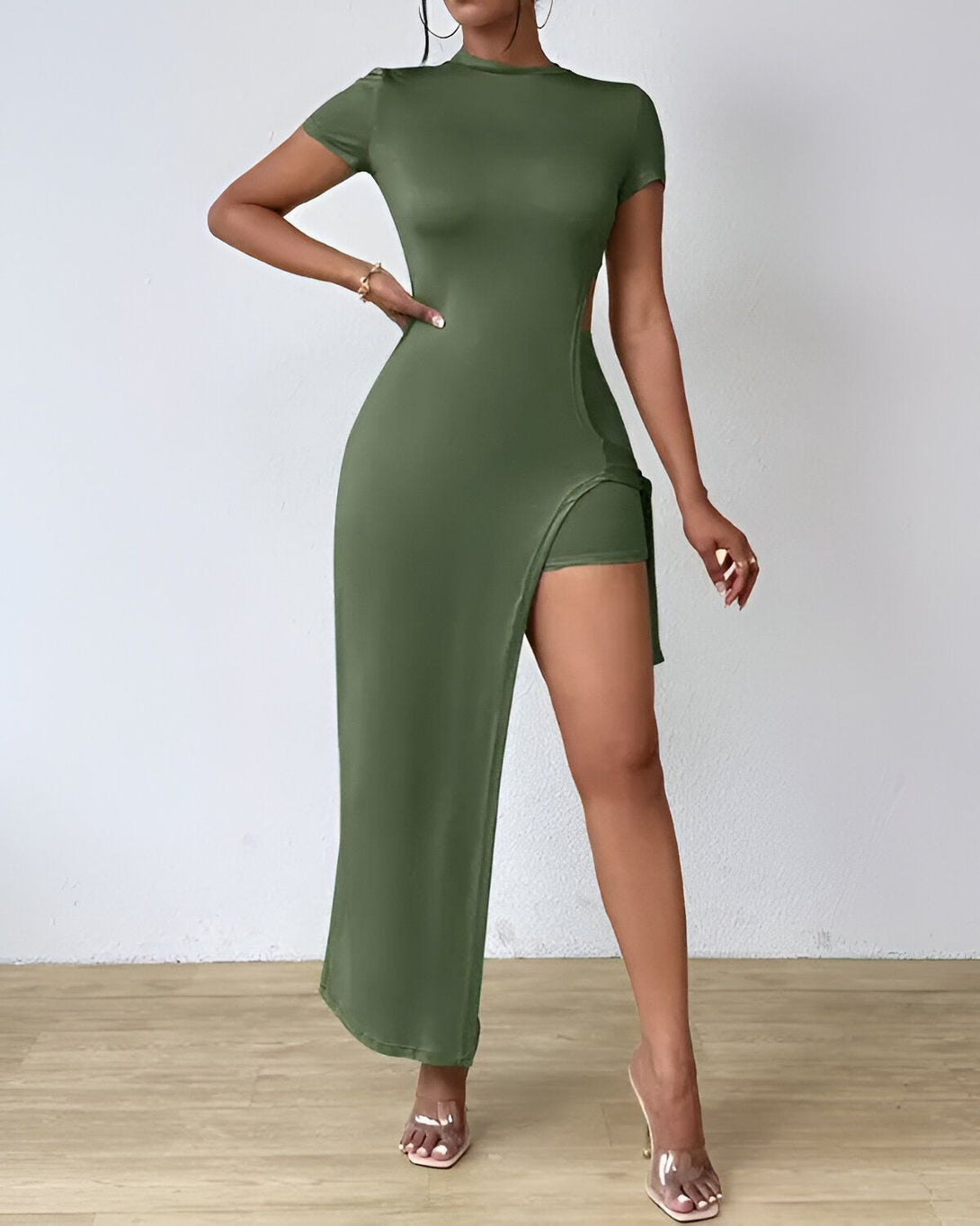 Une femme porte une robe longue verte fendue sur le côté avec un short intégré. Elle est assortie avec des sandales à talons transparentes.