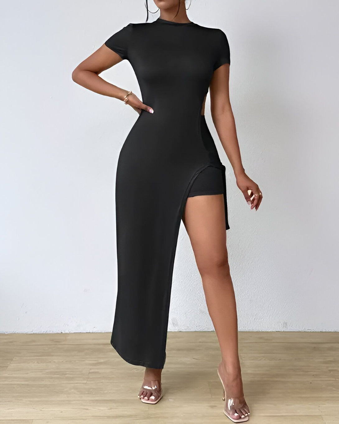 Une femme porte une robe noire moulante avec un shorty intégré. La robe est fendue sur le côté et elle porte des sandales à talons transparentes.