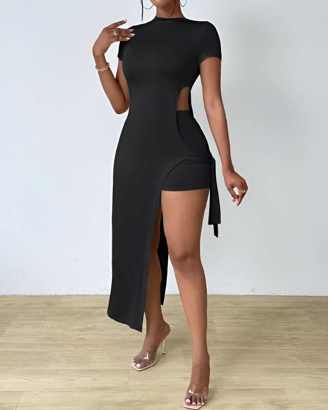 Une femme porte une robe noire moulante avec un shorty assorti. La robe a une fente sur le côté et le shorty est noué sur le côté. La femme porte des talons transparents et des bijoux dorés.