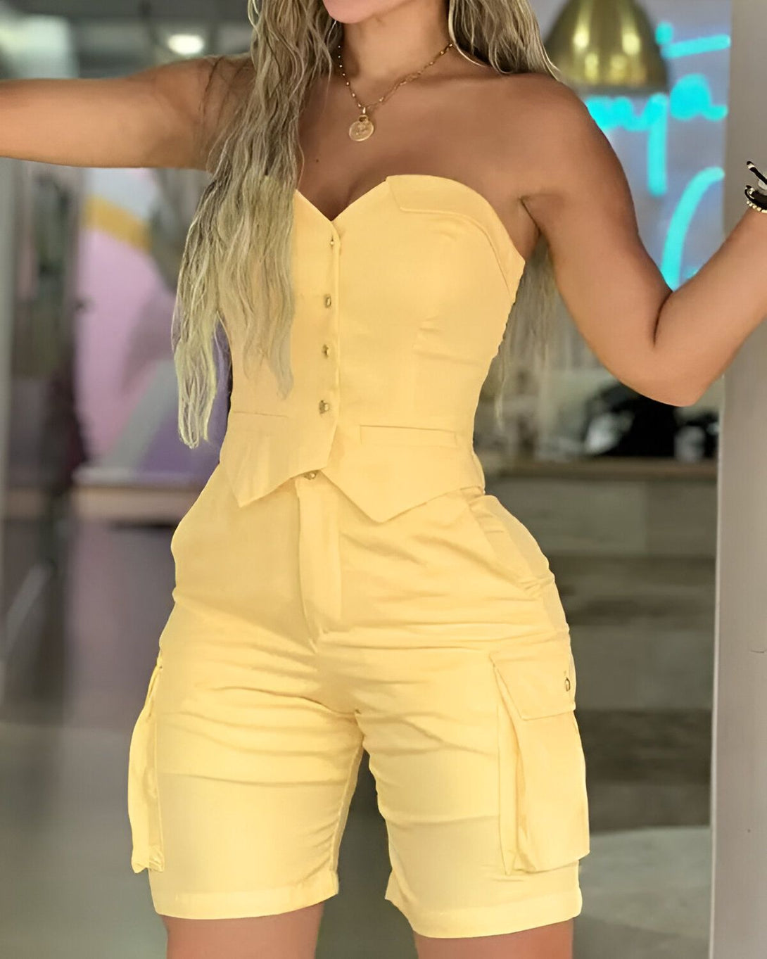 Une femme porte un ensemble jaune composé d'un bustier et d'un short cargo. Le bustier est strapless et possède des boutons sur le devant. Le short est taille haute et possède des poches cargo sur les côtés. La femme est blonde et porte un collier doré. Elle est debout dans une pièce et regarde vers la caméra.