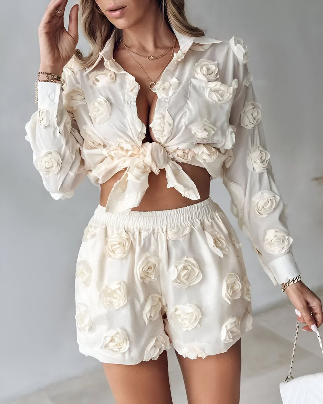 Une femme porte un ensemble deux pièces blanc cassé composé d'une chemise et d'un short. La chemise est ouverte sur le devant et nouée au niveau de la taille. Le short est taille haute et évasé. La femme est blonde et porte des talons hauts blancs. Elle est debout dans un intérieur et regarde l'appareil photo.