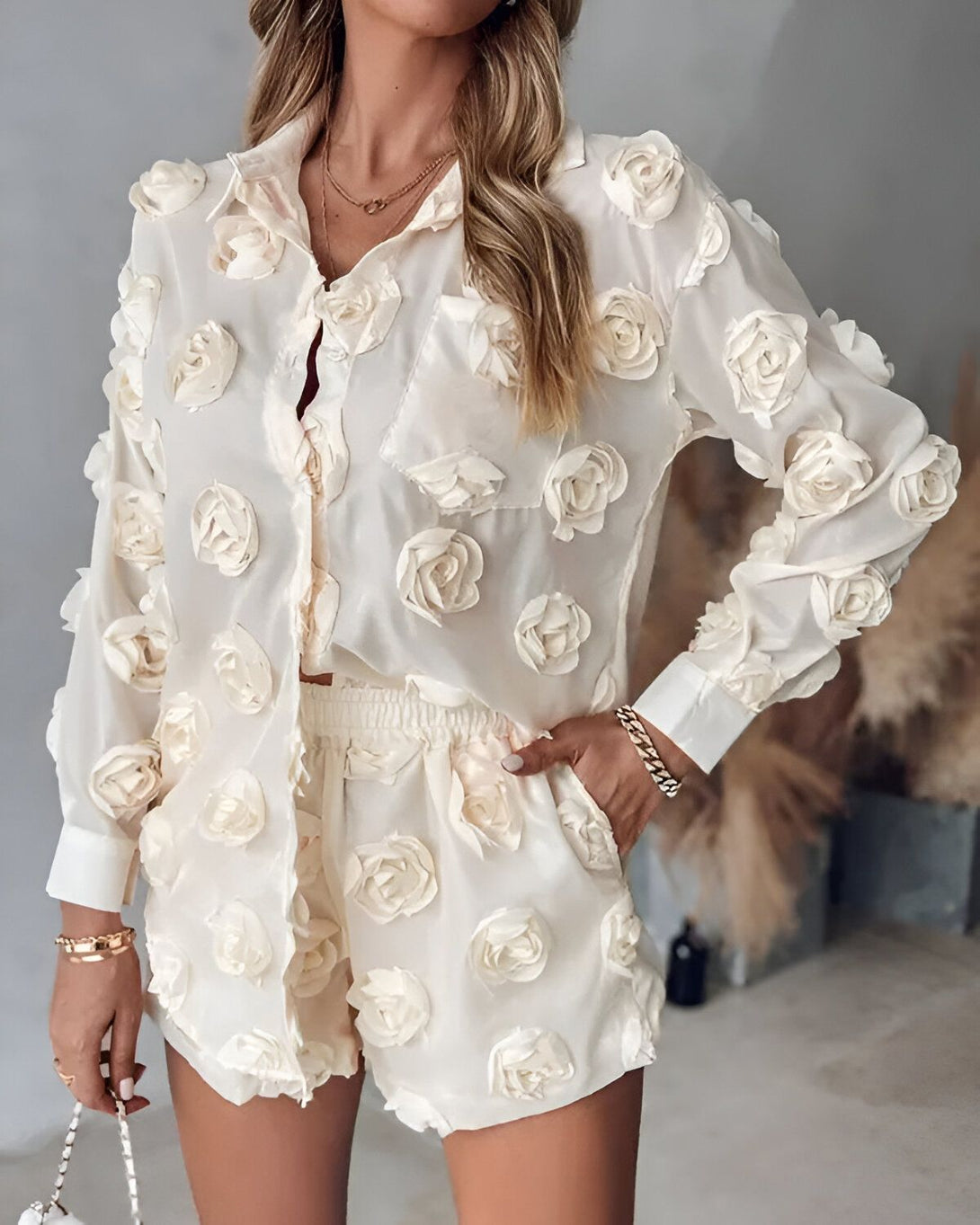 Une femme porte un ensemble short et chemisier blanc cassé. Le chemisier est ample et possède des manches longues. Il est entièrement recouvert de roses en relief de la même couleur. Le short est quant à lui taille haute et possède une ceinture élastique. Il est également recouvert de roses en relief. La femme porte des sandales à talons couleur chair et un collier doré. Elle a les cheveux longs et blonds. Elle est debout, les mains dans les poches du short. Derrière elle se trouve un mur beige.