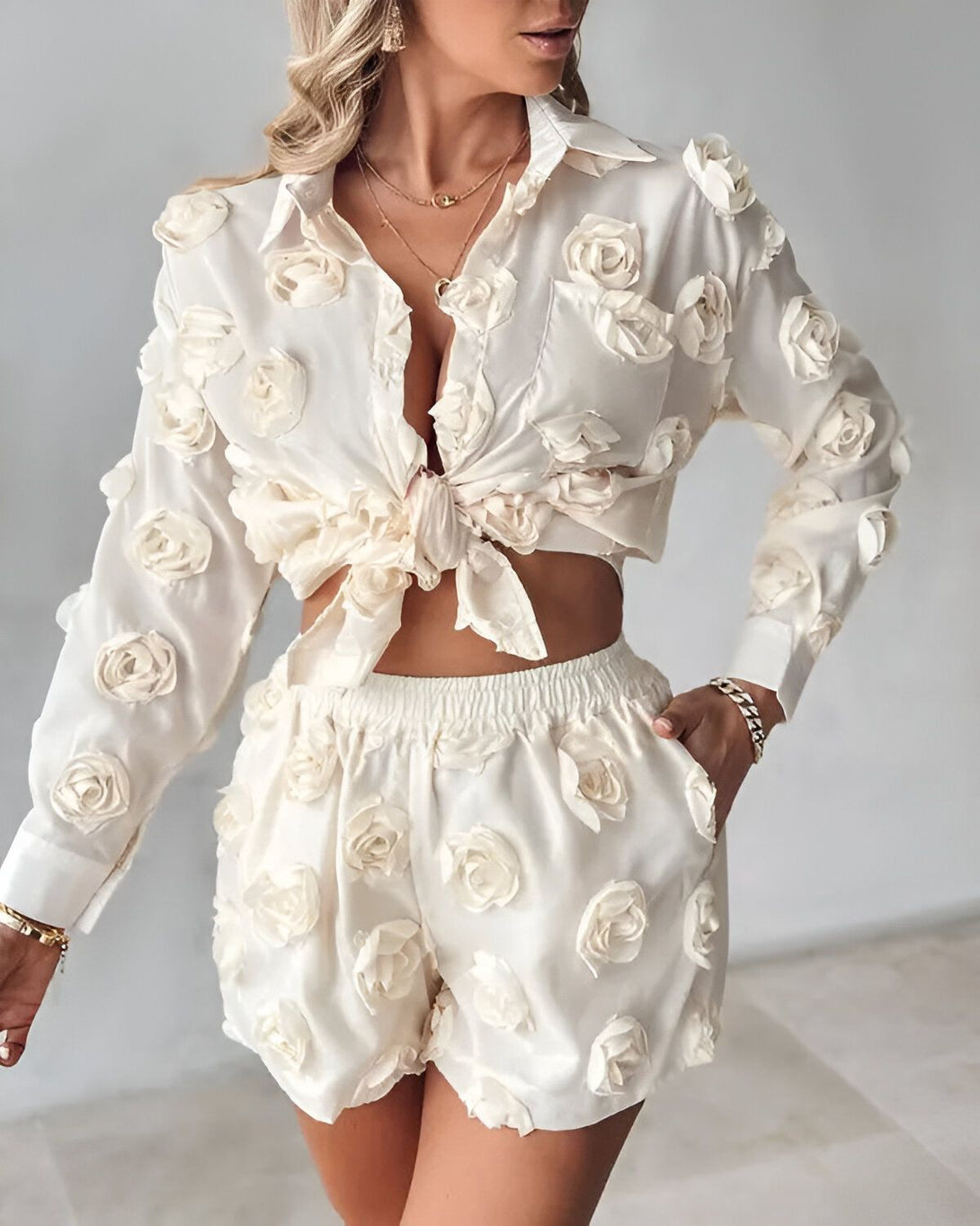 Une femme porte un ensemble short et chemisier blanc cassé. Le chemisier est noué au niveau du ventre et les manches sont bouffantes. Le short est taille haute et possède des poches.