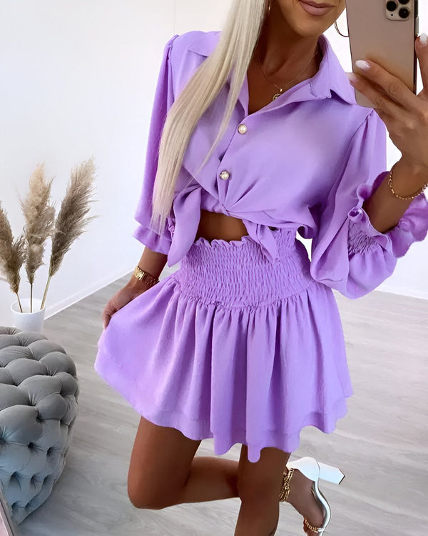 Une femme porte un ensemble jupe et chemisier lavande. La jupe est courte et évasée, avec une taille haute. Le chemisier est ample et noué au niveau de la taille. La femme est blonde et porte des talons blancs.