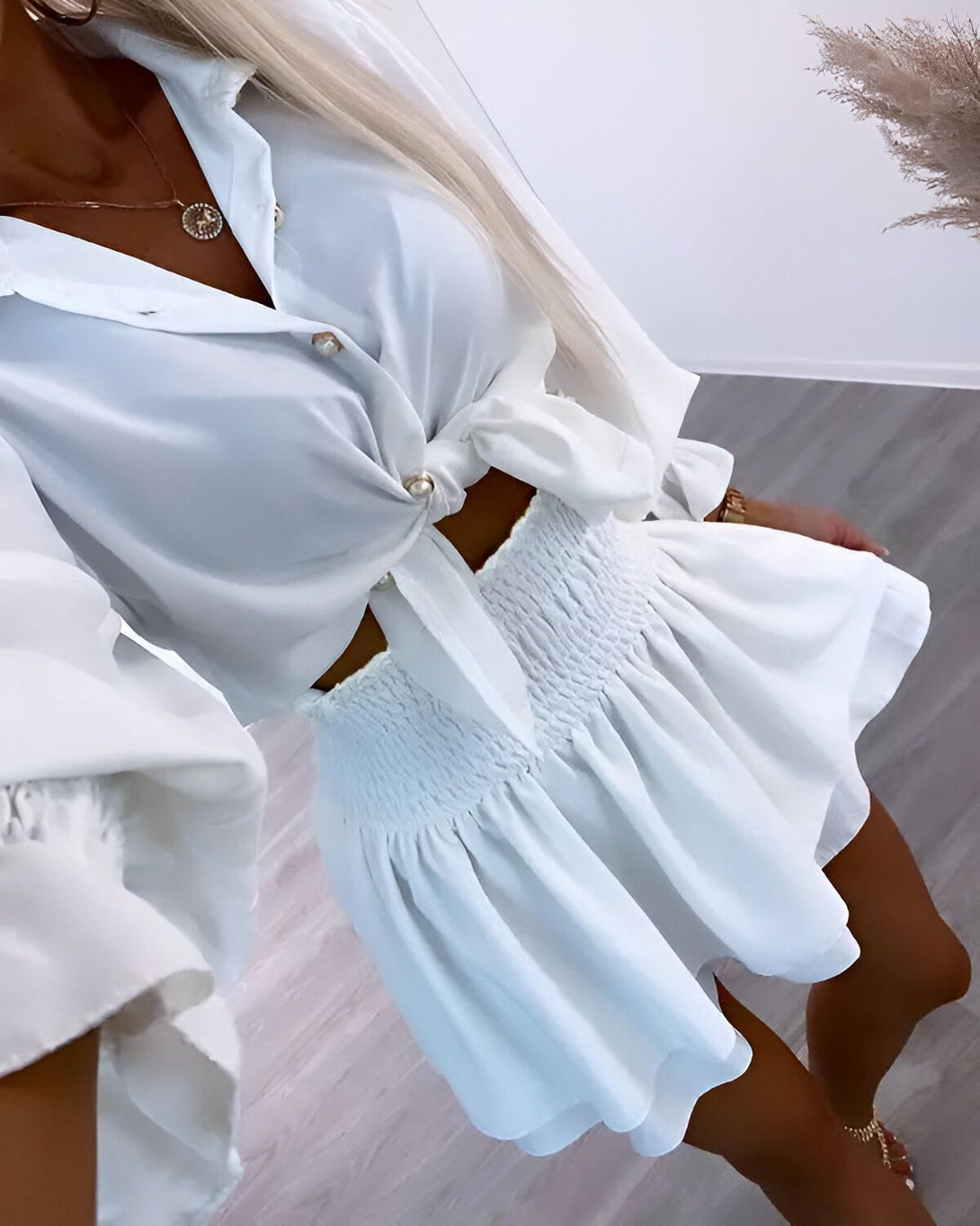 Une femme porte un ensemble blanc composé d'une chemise et d'une jupe. La chemise est ouverte et nouée au niveau du ventre. La jupe est courte et évasée. L'ensemble est complété par des sandales dorées.