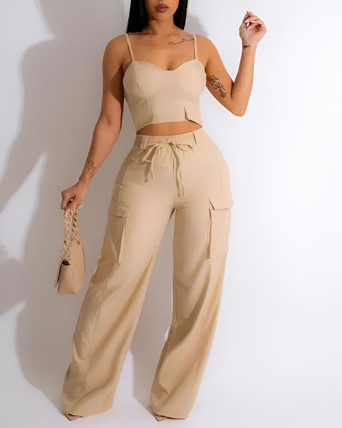 Une femme porte un ensemble beige composé d'un crop top et d'un pantalon cargo. Le crop top est moulant et possède des bonnets rembourrés. Le pantalon est ample et possède des poches cargo sur les côtés. La femme porte également un sac à main beige et des talons hauts.