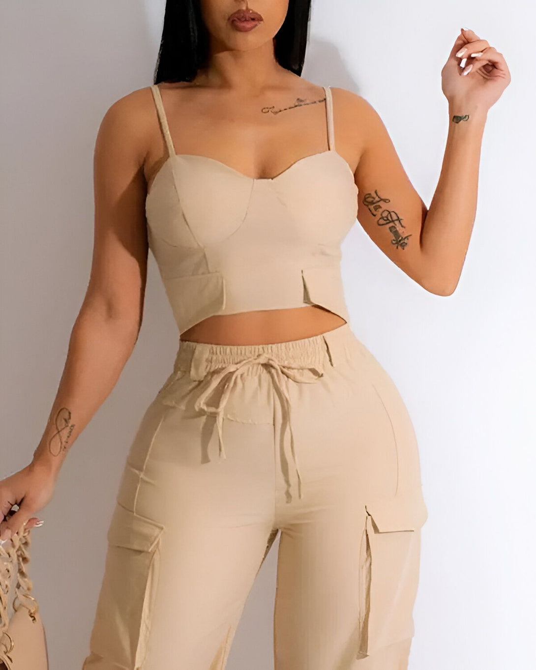 Une jeune femme porte un ensemble beige composé d'un crop top et d'un pantalon cargo. Le crop top est court et moulant, avec des bretelles fines. Le pantalon est ample et possède des poches cargo sur les côtés. La femme est debout, de face, et regarde l'appareil photo. Elle a les cheveux longs et noirs, et porte un maquillage léger.