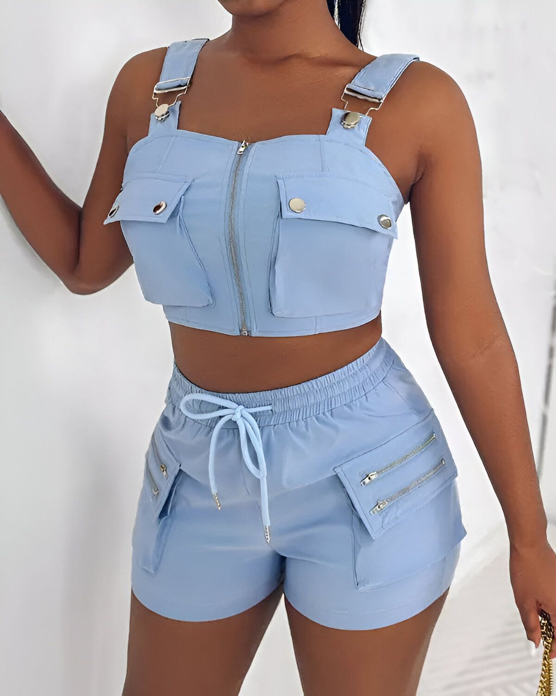 Une femme porte un ensemble deux pièces bleu clair composé d'un crop top et d'un short. Le crop top est zippé sur le devant et possède des poches sur le devant. Le short est taille haute et possède des poches zippées sur les côtés.