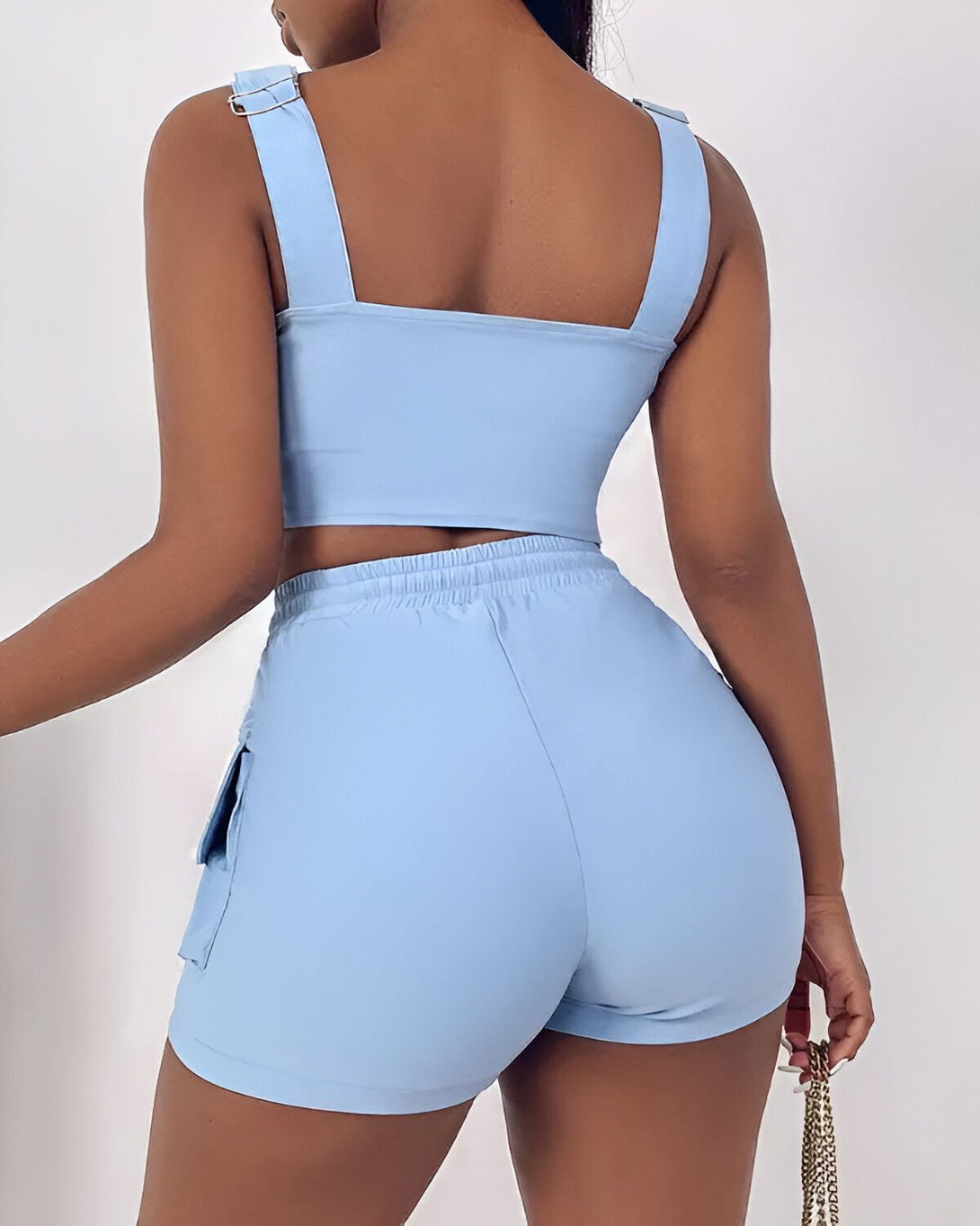 Une femme porte un ensemble de sport bleu ciel composé d'un crop top et d'un short. Le crop top est court et moulant, avec des bretelles fines. Le short est taille haute et possède des poches cargo sur les côtés. La femme est vue de dos et porte une chaîne en or autour du cou.