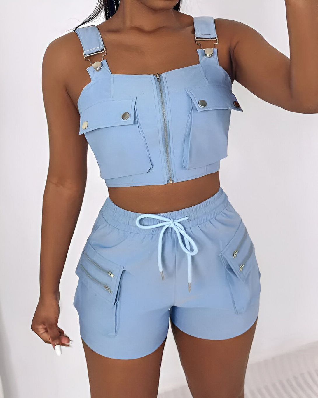 Une femme porte un ensemble deux pièces bleu clair composé d'un crop top et d'un short. Le crop top est zippé sur le devant et possède des poches sur le devant. Le short est taille haute et possède des poches zippées sur les côtés.