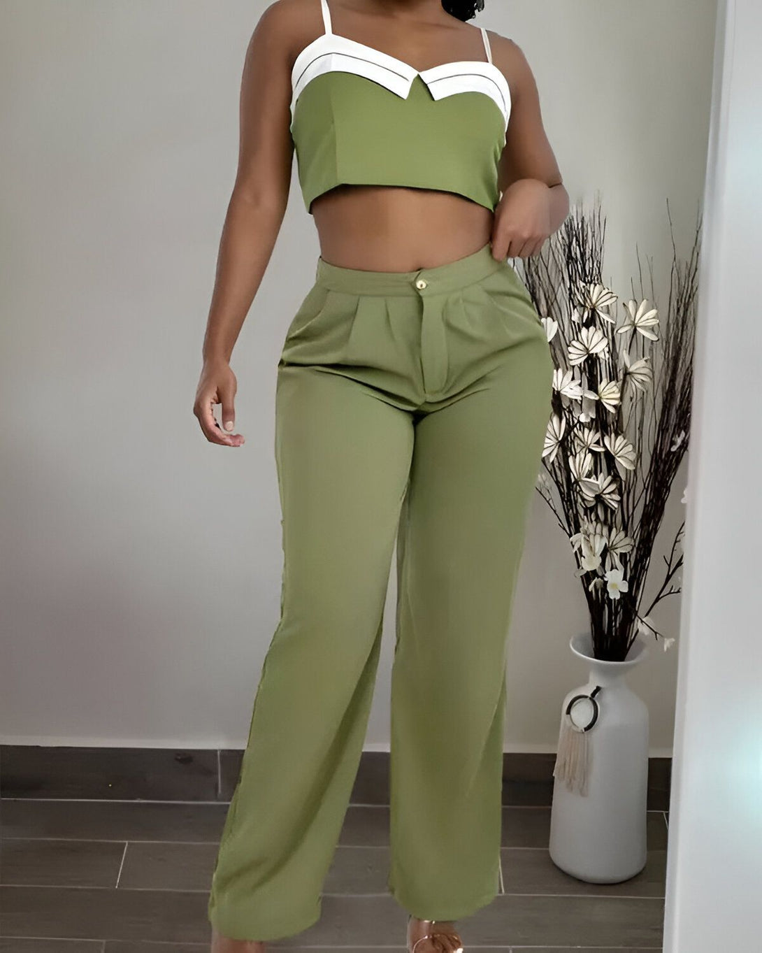 Une femme porte un pantalon taille haute vert olive et un crop top vert olive et blanc. Elle est debout, les mains sur les hanches, devant un mur blanc.