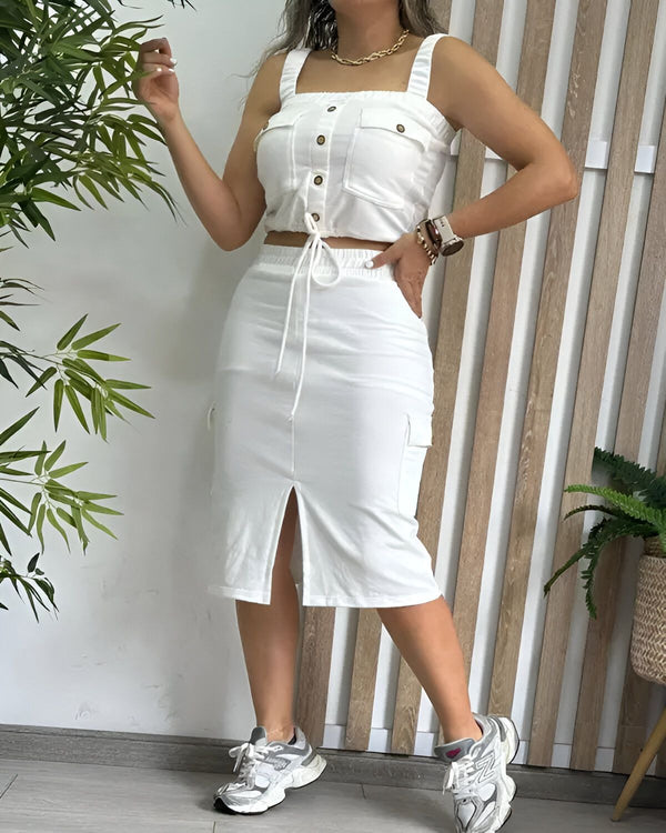 Une femme porte un crop top blanc et une jupe longue blanche. Le crop top est court et possède des boutons sur le devant. La jupe est fendue sur le côté et possède des poches cargo. La femme porte des baskets grises et des bijoux dorés. Elle est debout dans un environnement naturel avec des plantes vertes en arrière-plan.