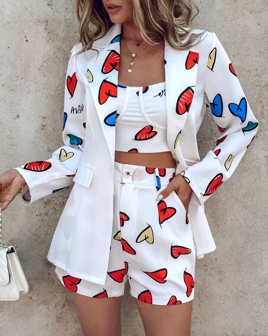 Une femme porte un tailleur blanc avec des motifs de cœurs colorés. Le tailleur est composé d'une veste et d'un short. La veste est ouverte et on peut voir un haut court rouge en dessous. Le short est taille haute et a des poches. La femme porte également un sac à main blanc.