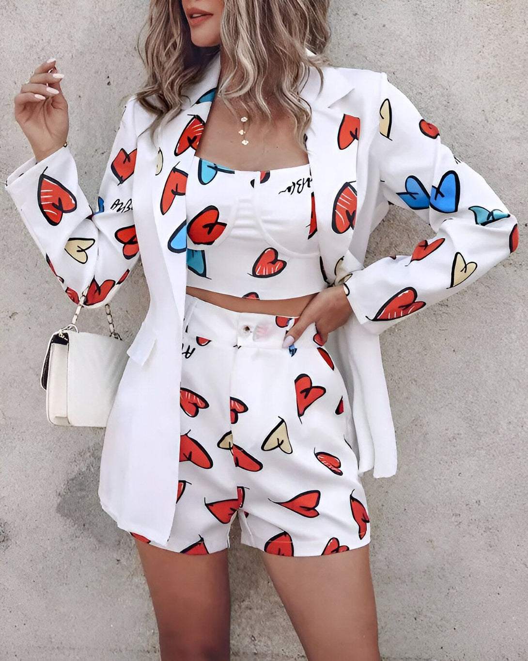 Une femme porte un tailleur blanc avec des motifs de cœurs colorés. Le tailleur est composé d'une veste et d'un short. La veste est ouverte et le short est taille haute. La femme porte également un top blanc. Elle a les cheveux longs et blonds. Elle est souriante et regarde l'objectif. Elle porte un sac à main blanc à la main droite.
