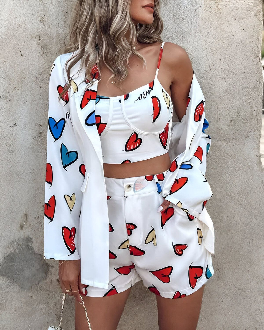 Une femme porte un blazer blanc à motifs de cœurs colorés, un crop top blanc à motifs de cœurs colorés et un short blanc à motifs de cœurs colorés.