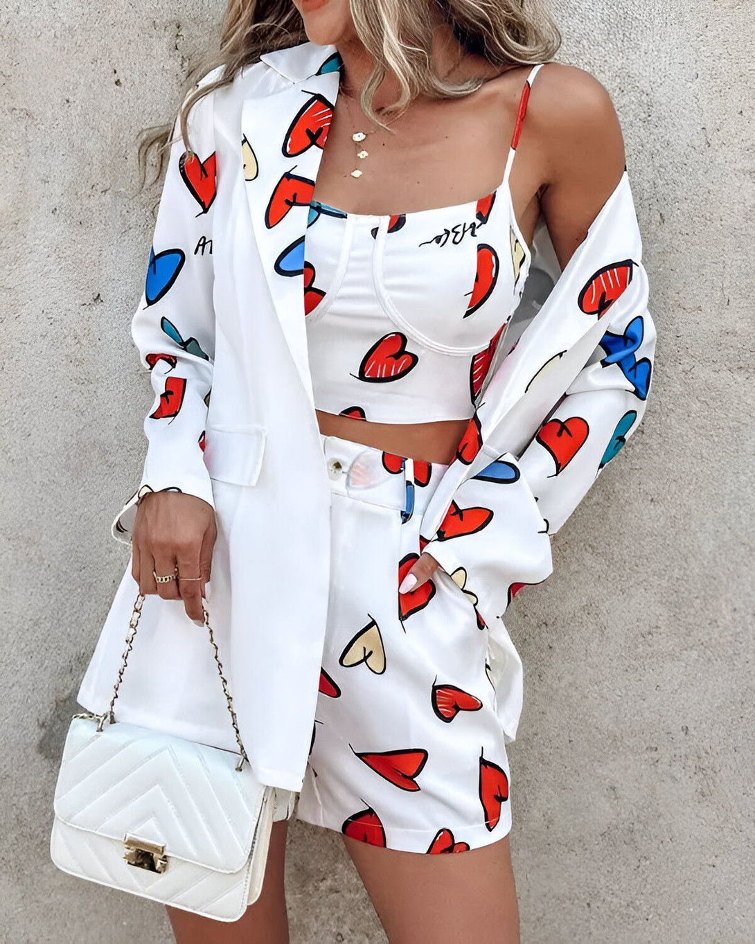 Une femme porte un blazer blanc à motifs de coeurs colorés, un crop top blanc et un short blanc assorti. Elle porte également un sac à main blanc.