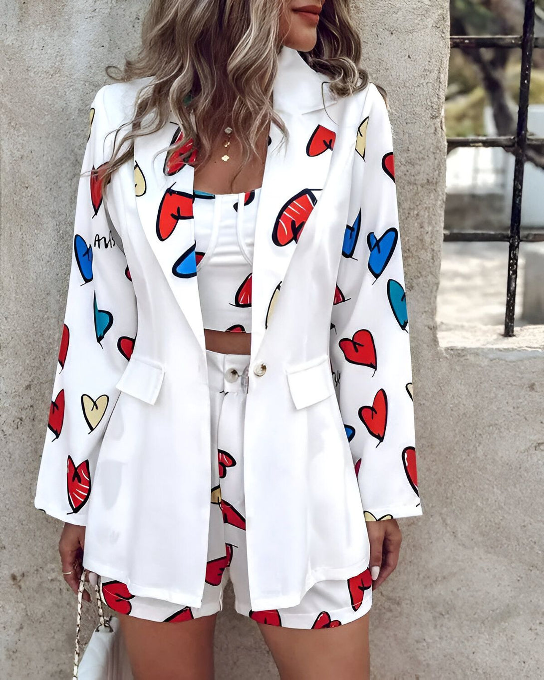 Une femme porte un tailleur blanc avec des motifs de coeurs colorés. Le tailleur est composé d'une veste et d'un short. La veste est ouverte et on peut voir un haut blanc en dessous. Le short est taille haute et ajusté. La femme porte des talons hauts blancs et un collier. Elle a les cheveux longs et blonds. Elle est debout devant un mur blanc.