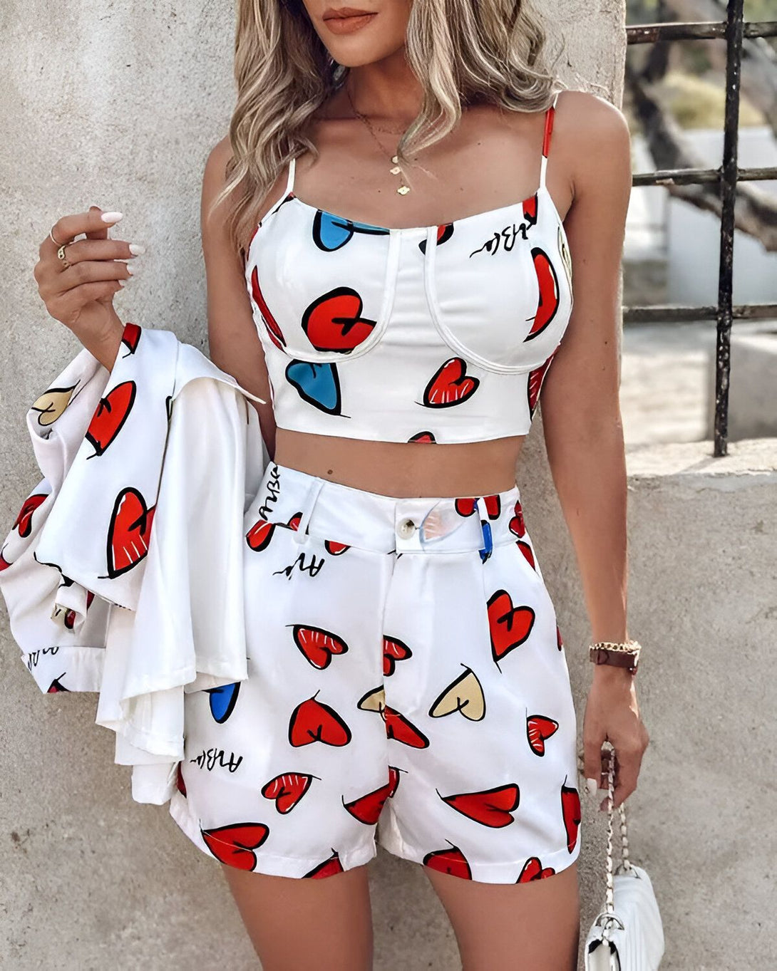 Une femme porte un crop top et un short assortis. Le crop top est blanc avec des cœurs rouges et bleus. Le short est blanc avec des cœurs rouges. La femme porte également une veste blanche.