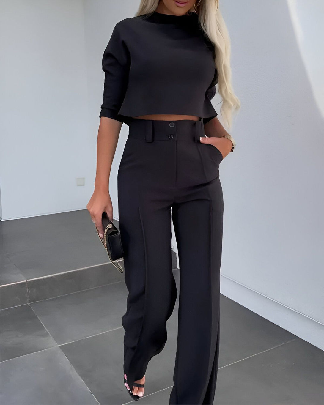 Une femme porte un tailleur pantalon noir avec un haut court et un pantalon large. Elle porte également des escarpins noirs et un sac à main noir.