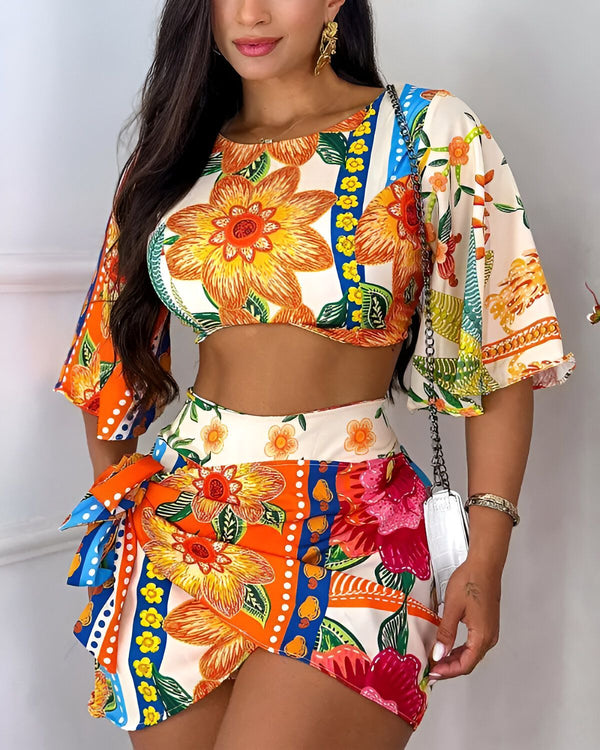 Une femme porte un ensemble deux pièces coloré avec un motif floral. Le haut est court et les manches sont bouffantes. Le bas est une jupe courte avec une fente sur le côté. La femme est debout et porte des talons hauts blancs. Elle a les cheveux longs et noirs et porte un maquillage léger.