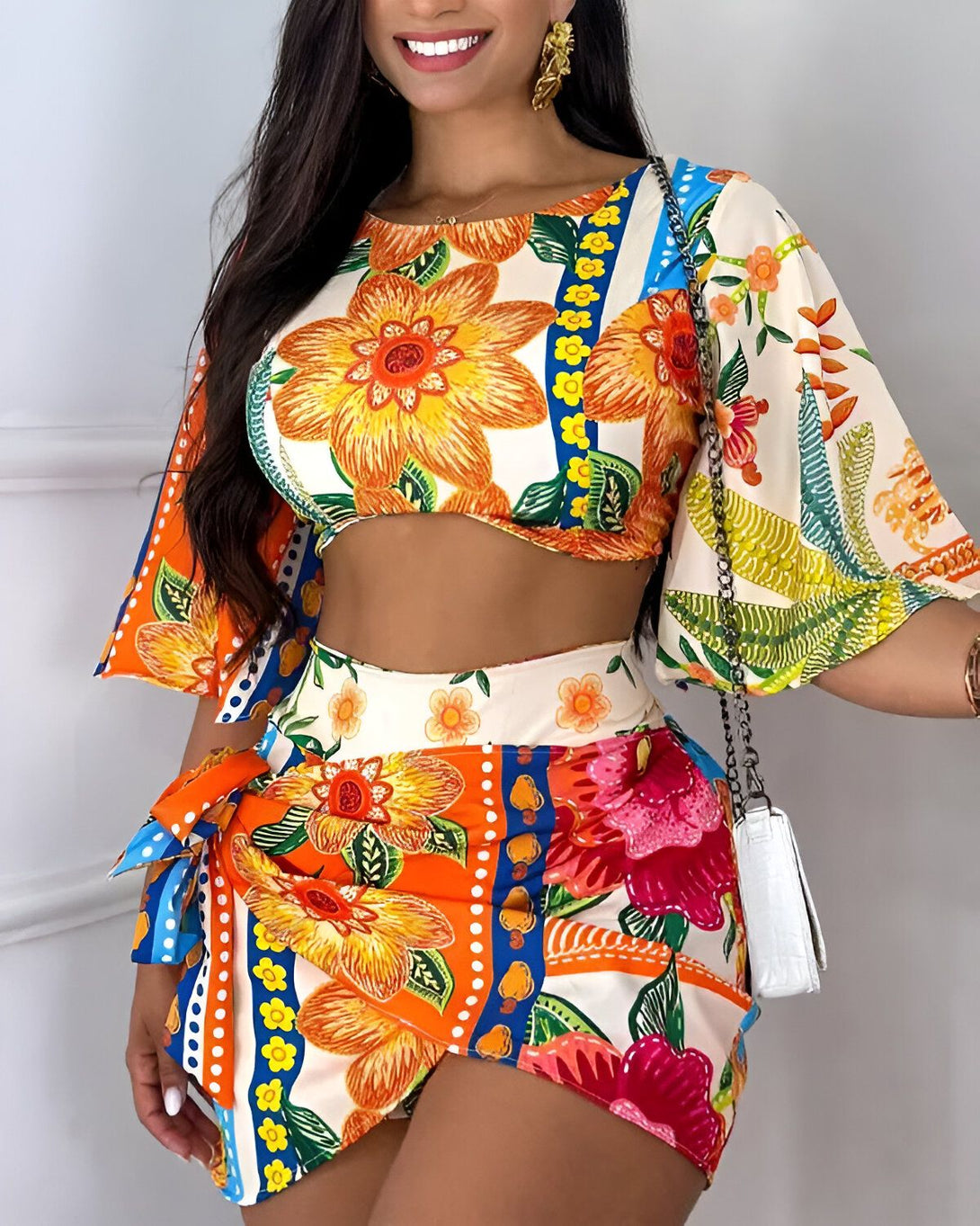 Une jeune femme souriante porte un ensemble deux pièces coloré à motifs floraux. Le haut est court et les manches sont bouffantes. Le bas est un short taille haute avec une ceinture à nouer.