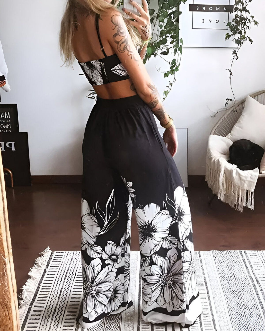 Une femme est debout dos à la caméra. Elle porte un haut court noir et blanc à motifs floraux et un pantalon large noir et blanc à motifs floraux. Elle est pieds nus et se trouve sur un tapis noir et blanc à motifs géométriques. En arrière-plan, on peut voir un mur blanc avec une plante verte et une chaise en osier sur laquelle est allongé un chat noir.