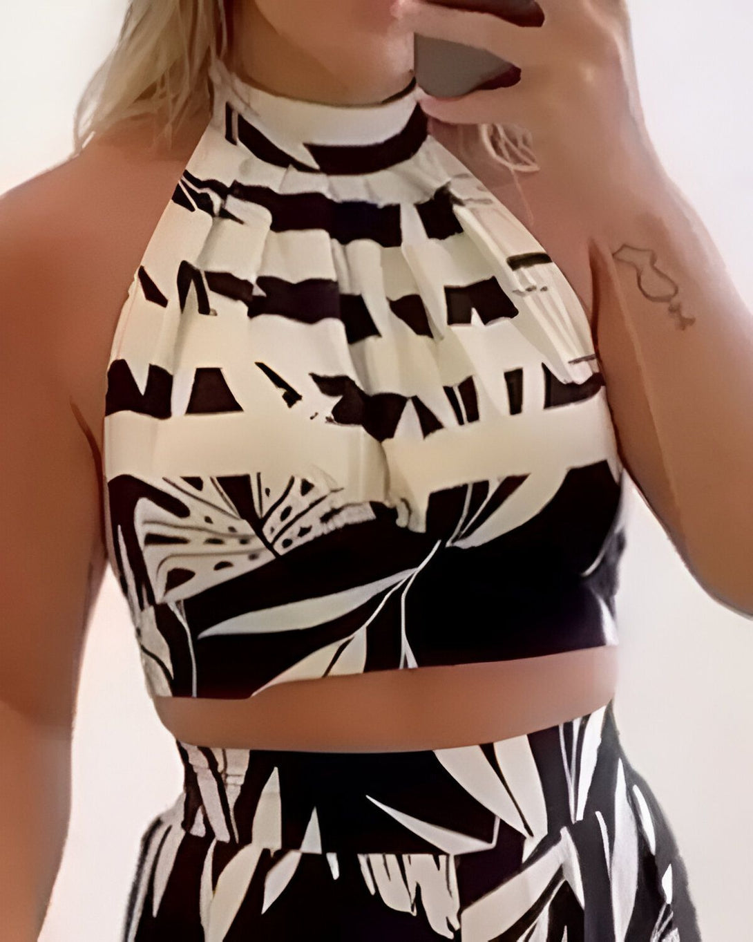 Une femme porte un haut de bikini noir et blanc à motifs de feuilles tropicales. Elle a les cheveux blonds et porte un collier ras du cou noir.