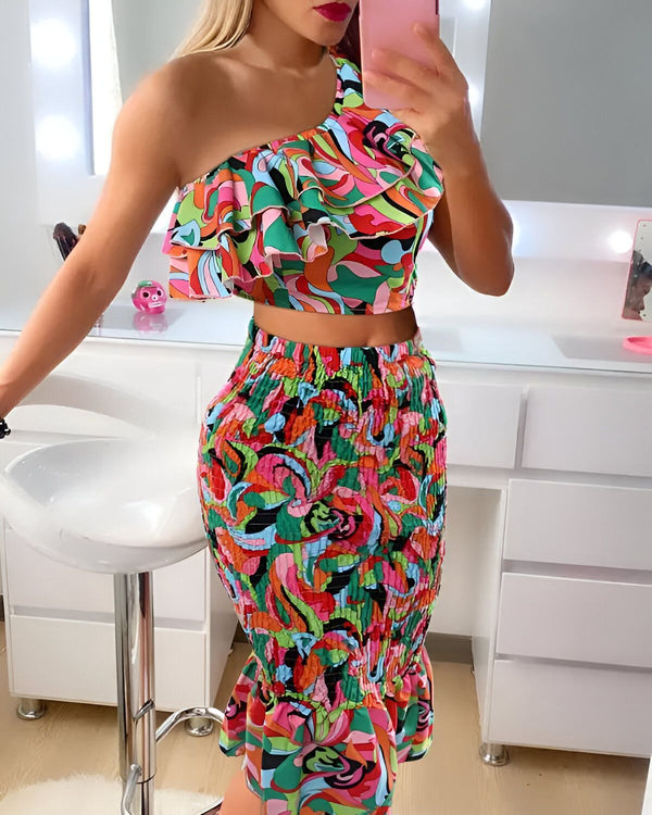 Une femme porte un ensemble coloré composé d'un crop top asymétrique et d'une jupe taille haute. Le crop top est à volants et l'ensemble est imprimé d'un motif floral. La femme est debout devant un miroir et se prend en photo avec son téléphone.