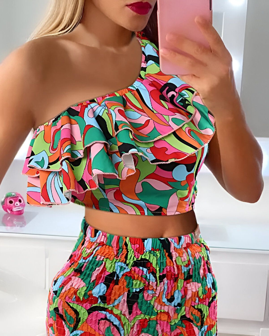 Une femme porte un haut court à une épaule et un pantalon assorti. Le haut est coloré et présente un motif psychédélique. Le pantalon est taille haute et a une coupe ample. La femme est blonde et a les lèvres rouges. Elle porte un maquillage léger et ses cheveux sont lâchés. Elle est debout dans une salle de bain et se prend en photo dans un miroir.