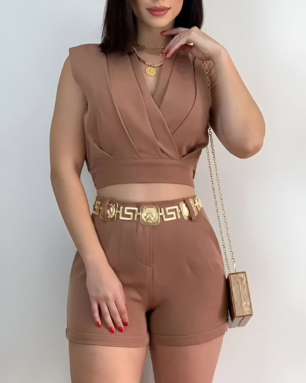 Une femme porte un ensemble pantalon et haut marron. Le haut est court et le pantalon est taille haute. Elle porte également une ceinture dorée et un collier doré. Elle a les ongles rouges et tient un sac à main transparent.