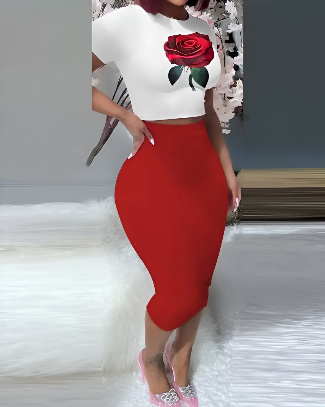 Une femme porte un crop top blanc avec une rose rouge imprimée dessus et une jupe crayon rouge. Elle porte également des talons aiguilles transparents.