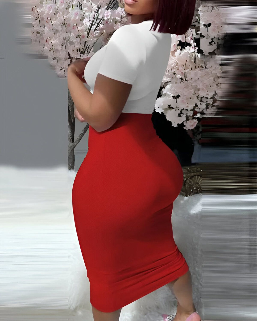Une femme porte une robe moulante rouge et blanche. La robe est courte et met en valeur ses courbes. Elle a des talons hauts blancs et des cheveux noirs. Elle est debout dans un salon, il y a un arbre avec des fleurs roses en arrière-plan.