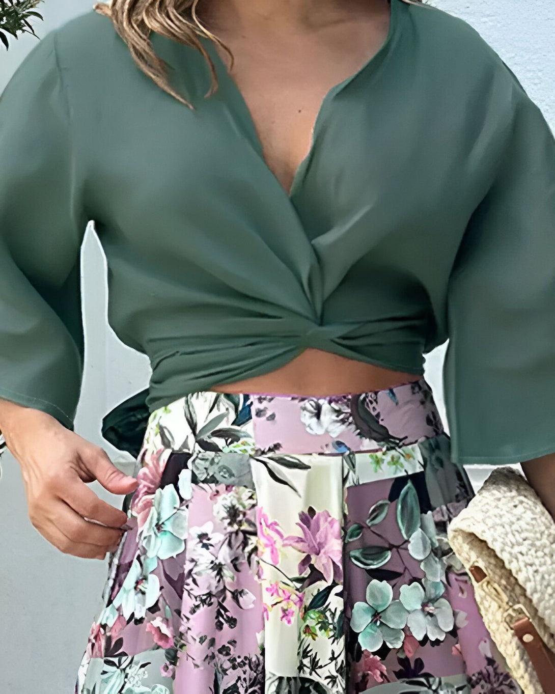 Une femme porte un chemisier vert et une jupe longue fleurie. Le chemisier est noué au niveau de la taille et la jupe est fendue sur le côté. La femme est debout, de face, et porte des sandales à talons. Elle a les cheveux longs et blonds et porte un maquillage léger.