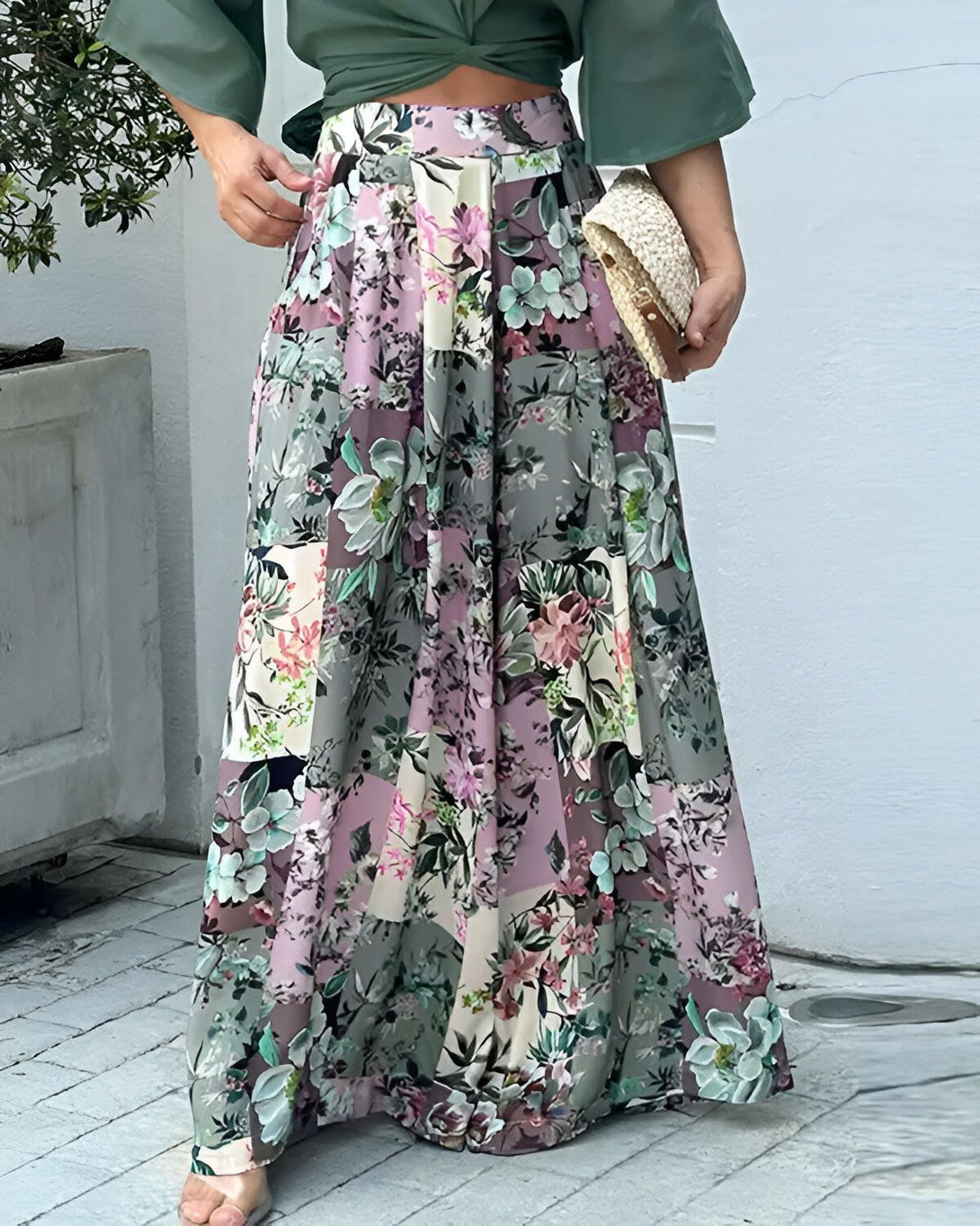 Une femme porte une jupe longue fleurie taille haute avec un chemisier vert noué sur le devant. Elle porte également un sac à main en paille et des sandales.
