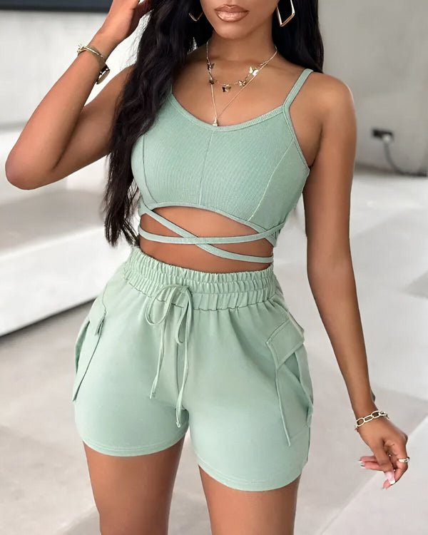 Une jeune femme porte un ensemble deux pièces vert sauge composé d'un crop top et d'un short. Le crop top est court et possède des bretelles fines. Il est croisé sur le devant et noué au niveau de la taille. Le short est taille haute et possède des poches cargo sur les côtés. Il est noué à la taille par un cordon de serrage. La femme est debout, les mains sur les hanches. Elle porte un collier et des boucles d'oreilles. Ses cheveux sont longs et noirs et elle est maquillée.