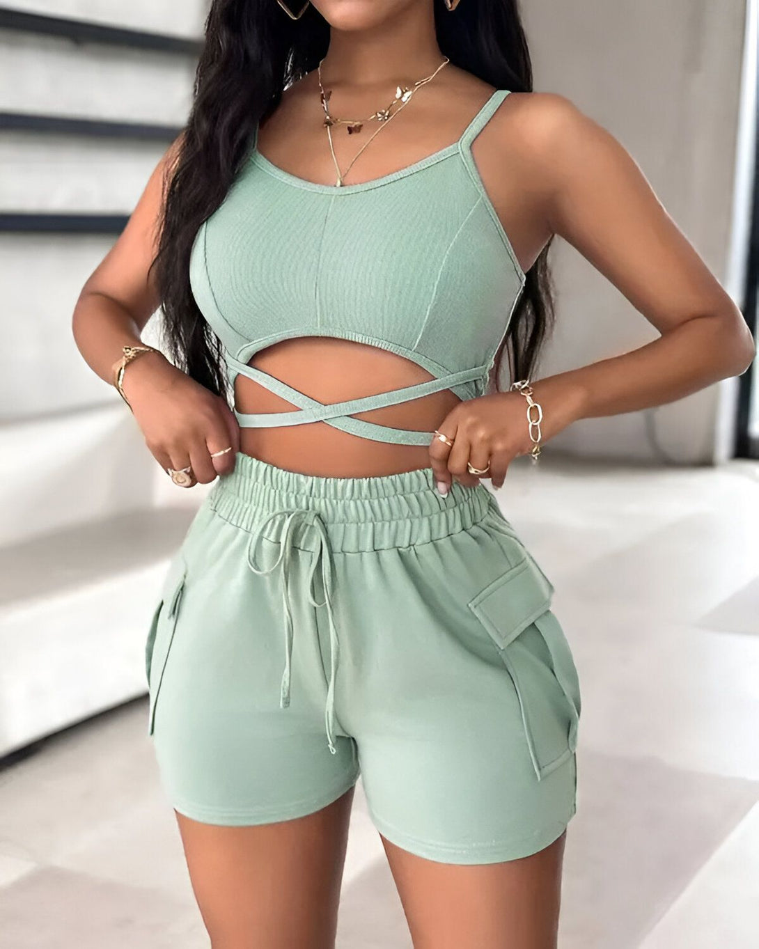 Une femme porte un ensemble deux pièces vert composé d'un crop top et d'un short. Le crop top est court et moulant, avec des bretelles fines et un décolleté croisé. Le short est taille haute et ample, avec des poches cargo sur les côtés. La femme est debout, les mains sur les hanches, et regarde l'appareil photo.