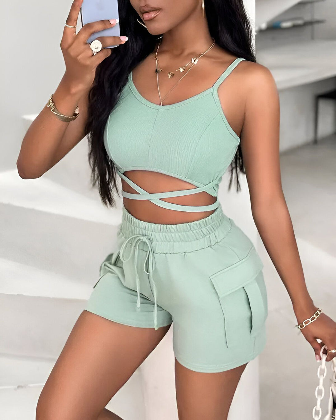 Une jeune femme porte un ensemble deux pièces vert composé d'un crop top et d'un short. Le crop top est court et possède des bretelles fines. Il est noué au niveau du ventre. Le short est taille haute et possède des poches cargo. La femme est debout et prend un selfie. Elle porte des bijoux et a les cheveux longs et noirs.