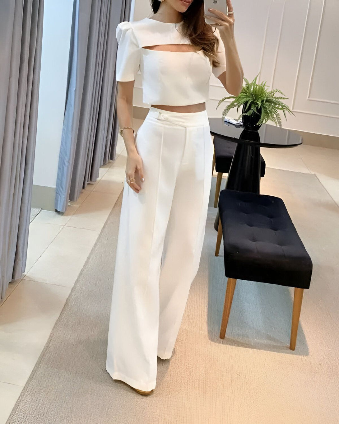 Une femme porte un tailleur pantalon blanc cassé. Le pantalon est large et taille haute. Le haut est court et laisse entrevoir un décolleté.