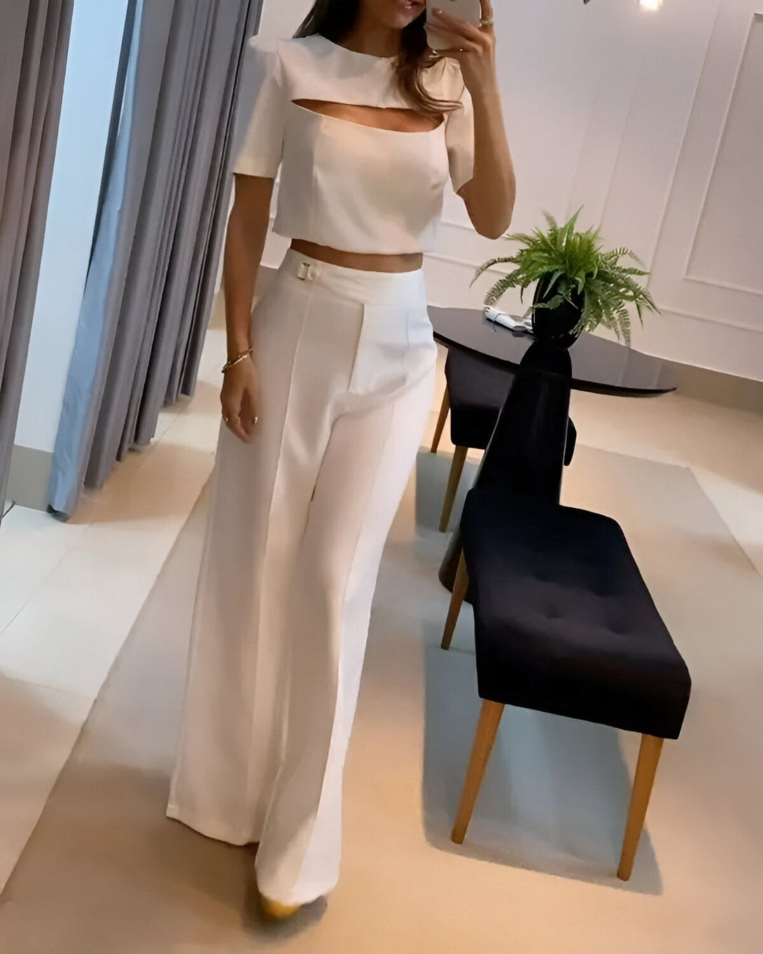 Une femme porte un pantalon taille haute blanc et un crop top blanc. Elle est debout dans un salon et se prend en photo dans un miroir.