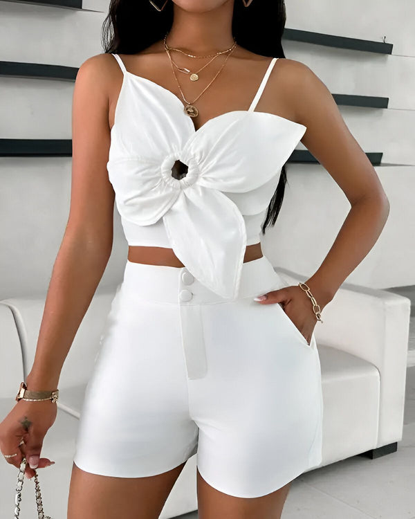 Une femme porte un ensemble deux pièces blanc composé d'un crop top et d'un short. Le crop top est doté de fines bretelles et d'un nœud papillon sur le devant. Le short est taille haute et possède des poches latérales. La femme est également porte une paire de sandales à talons blanches et une montre.