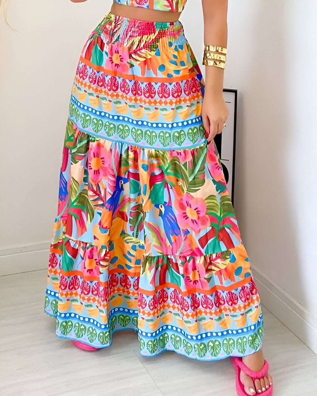 Une femme porte une longue jupe colorée à motifs tropicaux. La jupe est taille haute et évasée. Elle est composée de plusieurs volants. La femme est également porte un crop top blanc. Elle est pieds nus.