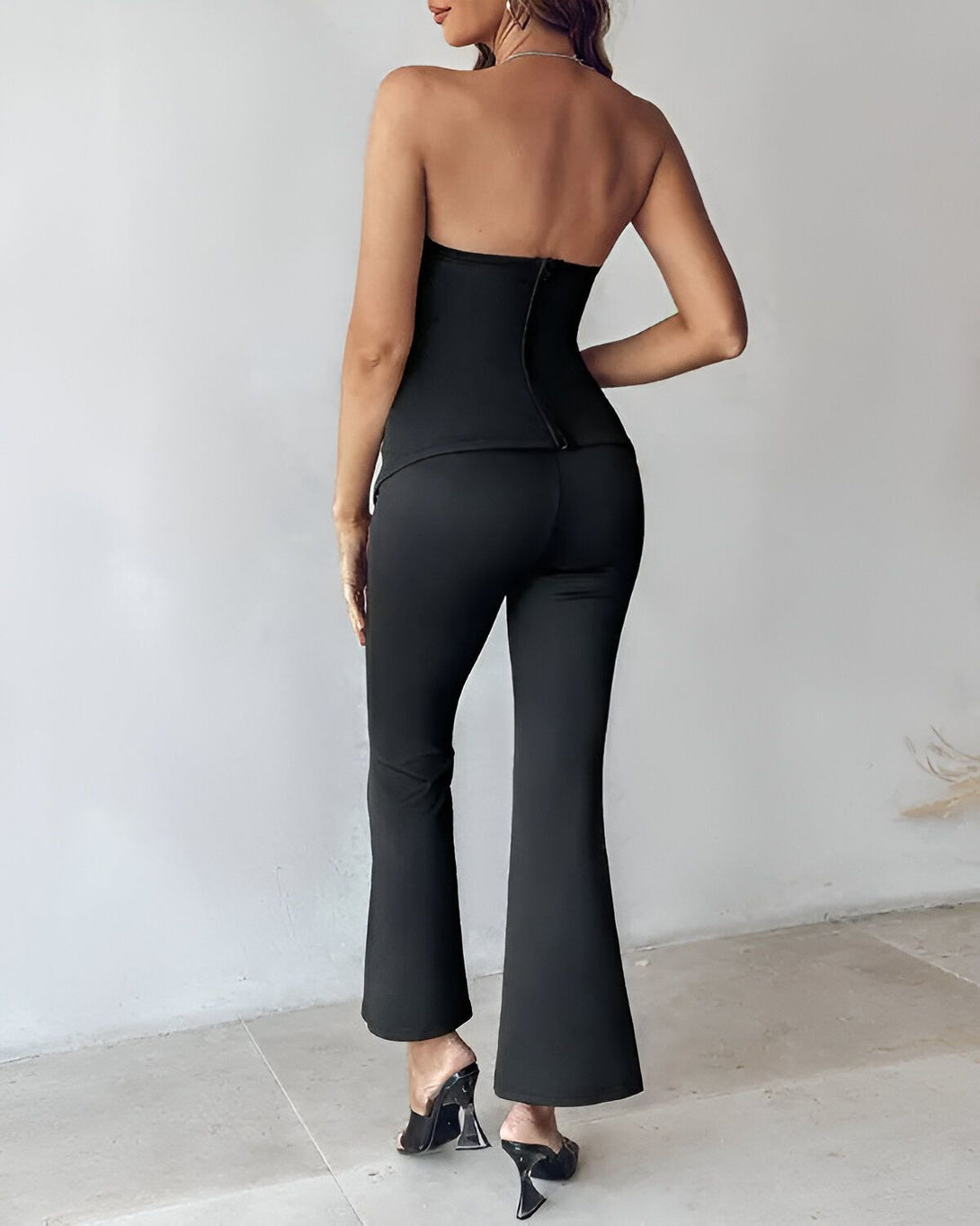 Une femme porte un bustier noir et un pantalon évasé noir. Elle est vue de dos.