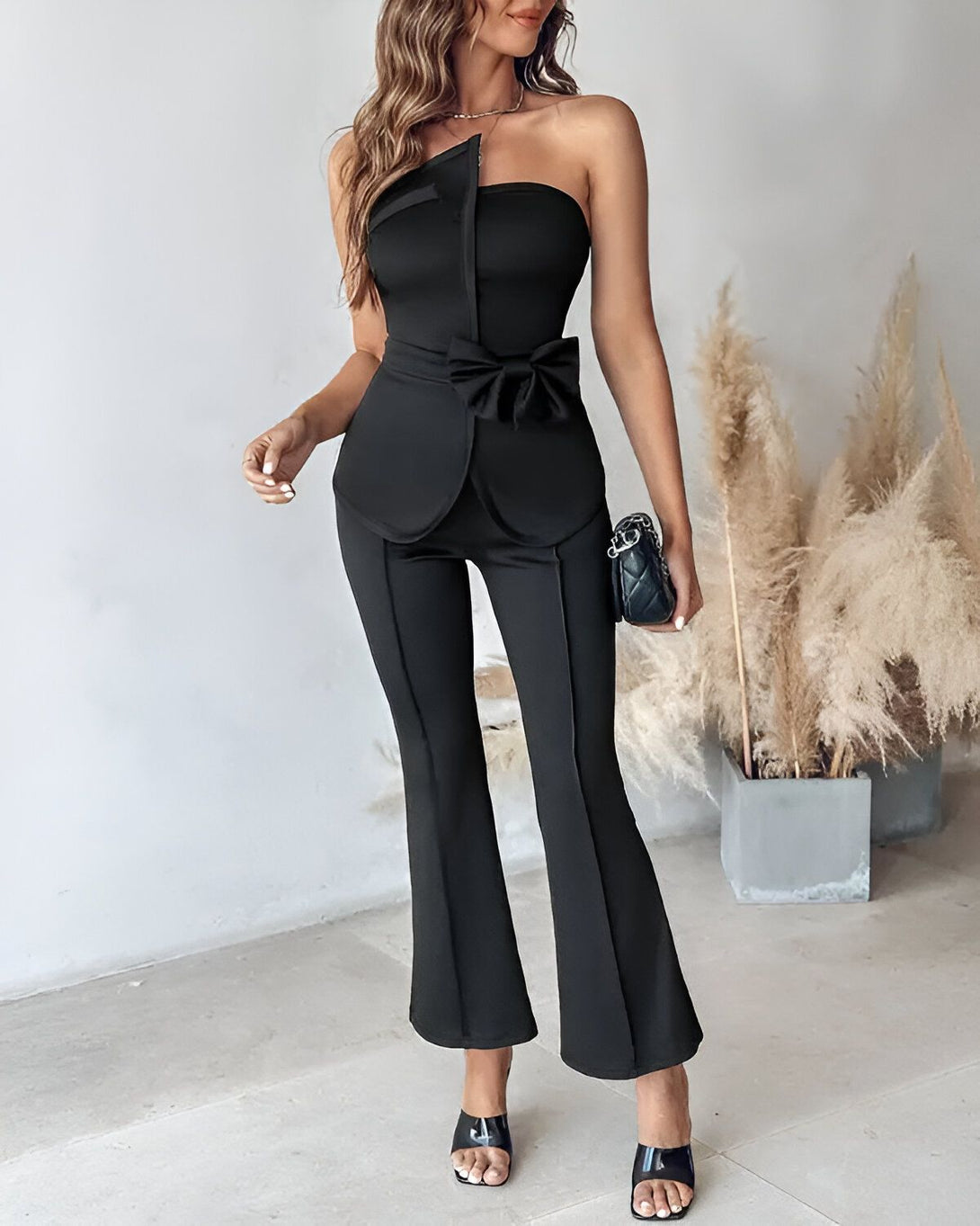 Une femme porte un ensemble pantalon et veste noir. Le pantalon est évasé et la veste est cintrée avec un nœud sur le devant. Elle porte des sandales à talons noires et un collier.