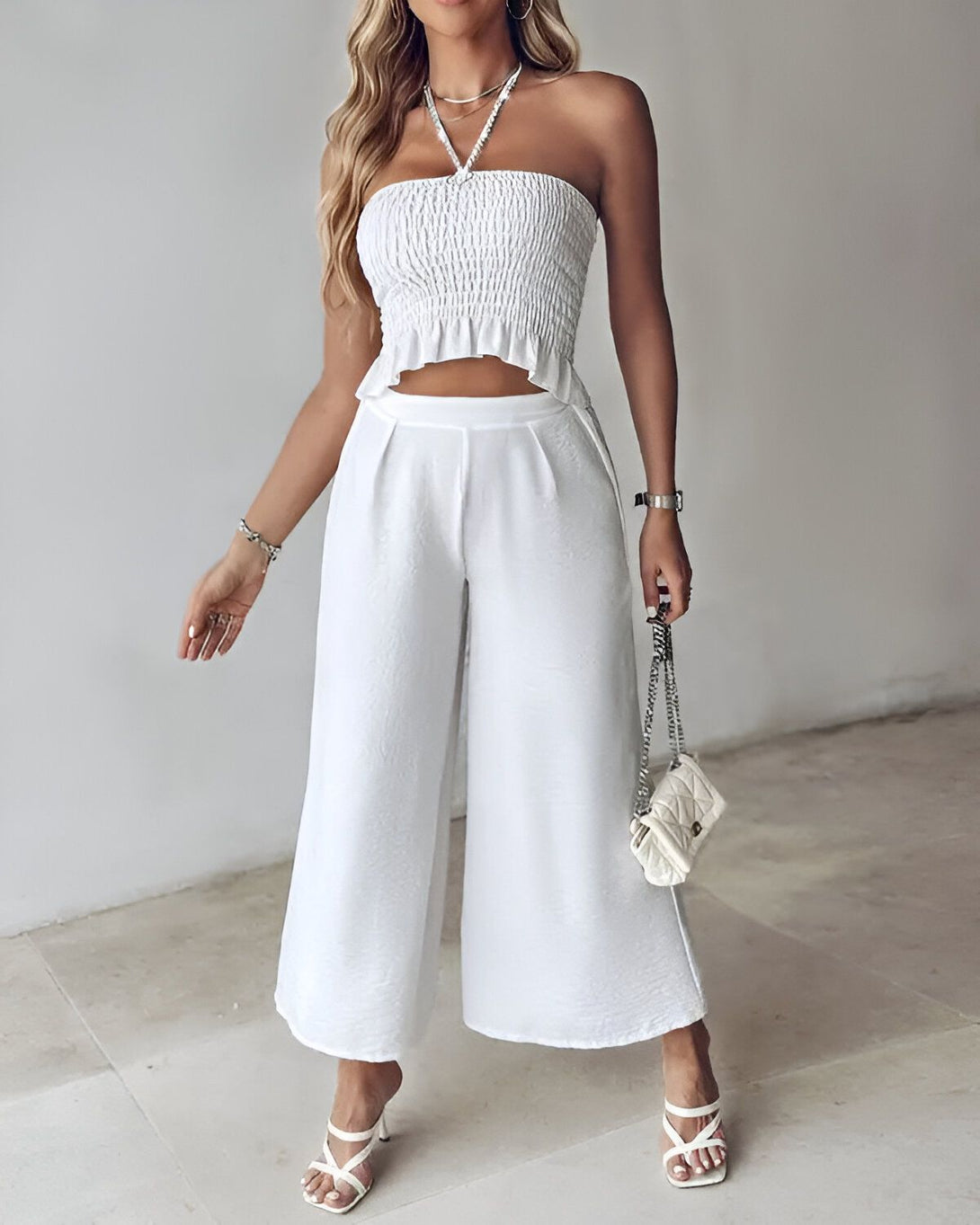 Une femme porte un haut court blanc et un pantalon large blanc. Elle porte également des sandales à talons blancs et un collier. Elle a un sac à main blanc à la main.