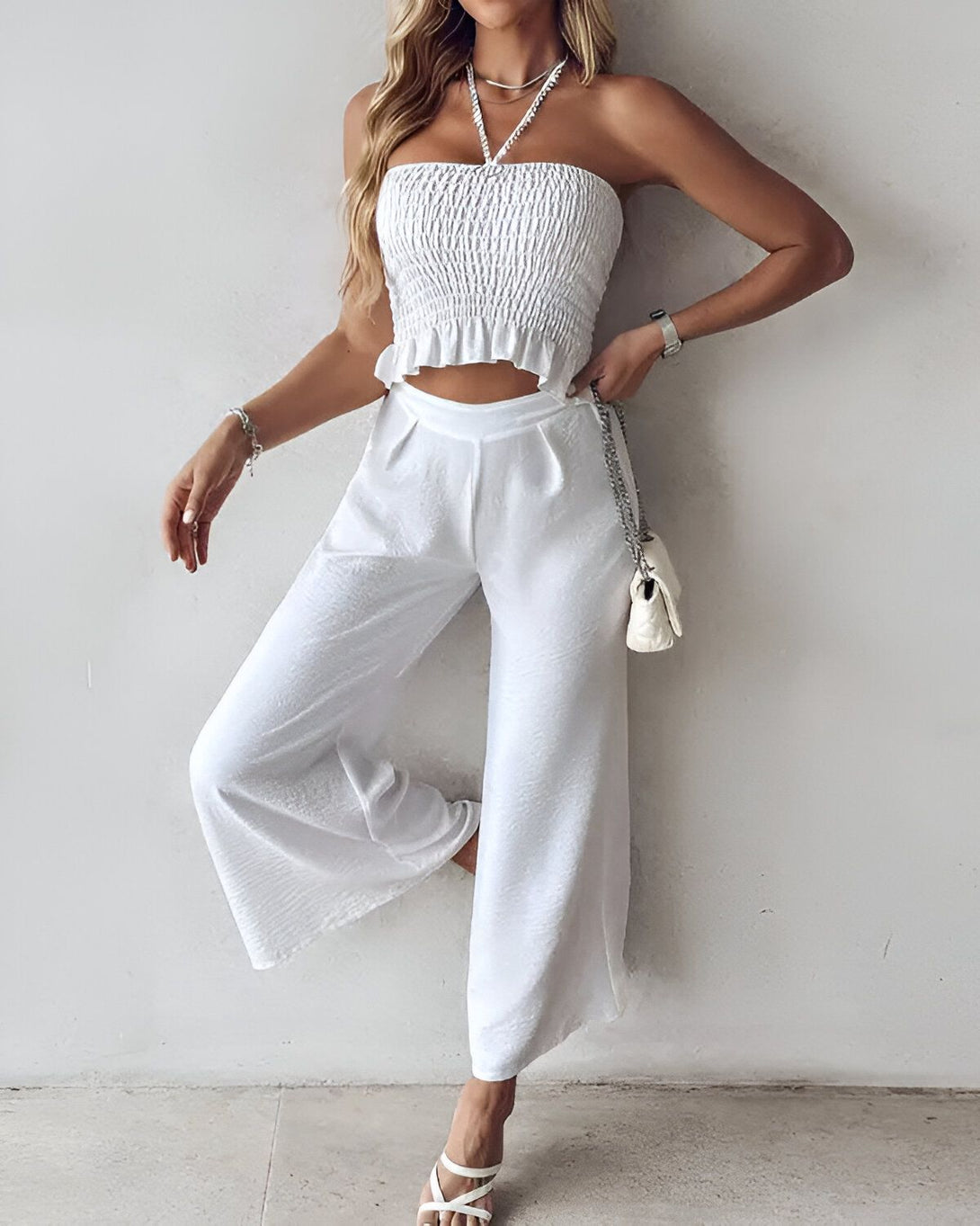 Une femme porte un haut court blanc et un pantalon large blanc. Elle est également porte des sandales à talons blancs et un collier.