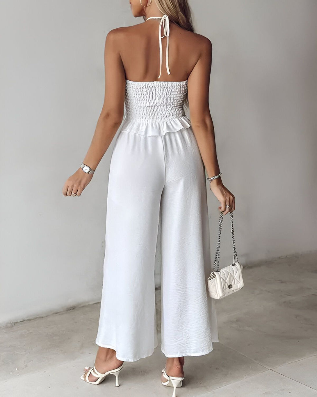 Une femme porte un pantalon et un haut blancs. Le pantalon est large et le haut est court. Elle porte également des sandales à talons hauts et un sac à main blanc.