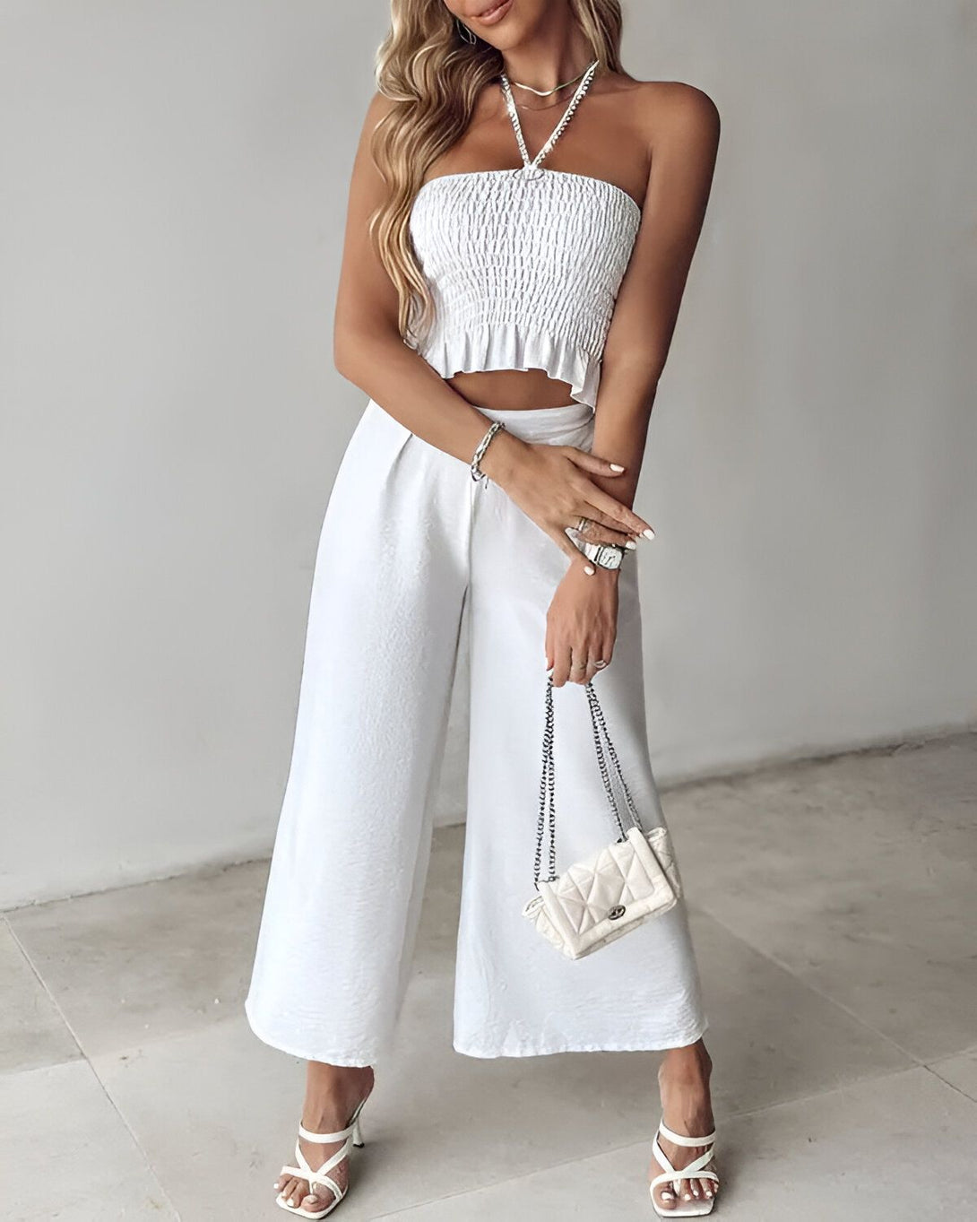 Une femme porte un haut court blanc et un pantalon large blanc. Elle porte également des sandales blanches et un collier. Elle tient un sac à main blanc à la main.