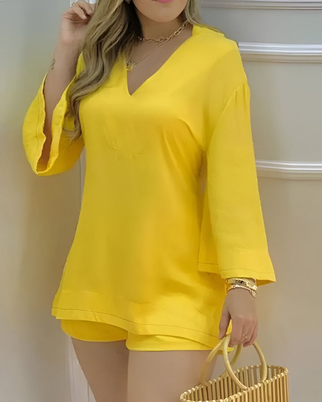 Une femme porte un ensemble short et haut jaune. Le haut est ample et possède des manches 3/4. Le short est taille haute. La femme est blonde et porte un collier doré. Elle est debout, de face, et tient un sac à main en osier à la main droite.