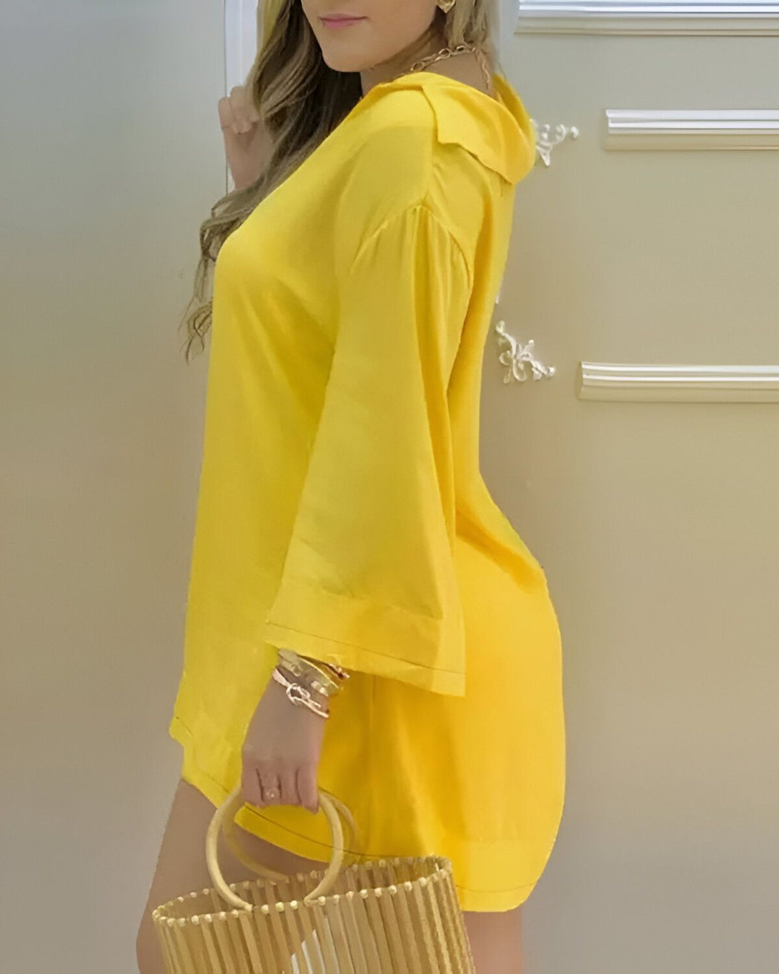 Une femme porte une robe jaune ample avec des manches bouffantes. Elle a les cheveux longs et blonds et porte un sac à main en osier à la main.